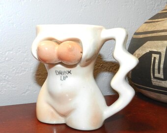 May include: A novelty ceramic mug shaped like a torso, with a white body and peach-colored breasts. The mug has a zig-zag handle and the words "DRINK UP" printed on the front. The mug is on a brown surface.