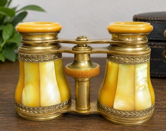 May include: Antique opera glasses with a brass frame and yellow and white mother-of-pearl panels. The lenses are framed with a decorative brass trim. The eyepieces are a light amber color. A vintage accessory.
