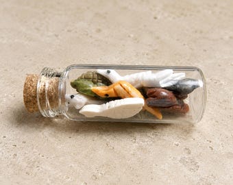 May include: A clear glass bottle with a cork stopper, filled with small, colorful, sculpted sea creatures. The creatures are various colors, including white, orange, green, and brown. The bottle is resting on a light-colored surface.
