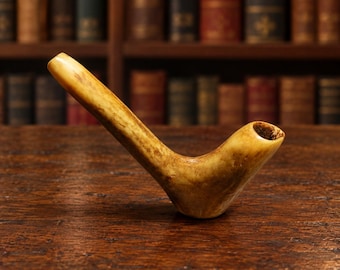 May include: A tobacco pipe crafted from a single piece of light tan, textured material with a smooth, polished finish. The pipe features a long, angled stem leading to a rounded bowl with a dark, hollow interior opening at the top end.