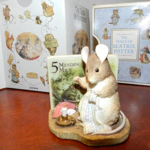 May include: A figurine of a mouse from Beatrix Potter's "The World of Beatrix Potter" series. The mouse is holding a book titled "5 Mending Mice" and is dressed in a white apron. The figurine is on a wooden base, with a candle and a book in the background.