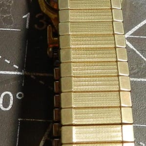 May include: A gold-tone metal watch band featuring a flexible, expandable link design with a textured, herringbone-style pattern on each rectangular segment. The band is attached to a watch case with a dark, smooth back, shown in a vertical orientation.