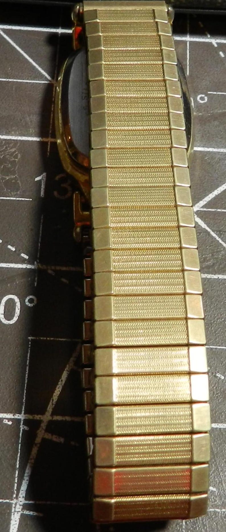 May include: A gold-tone metal watch band featuring a flexible, expandable link design with a textured, herringbone-style pattern on each rectangular segment. The band is attached to a watch case with a dark, smooth back, shown in a vertical orientation.