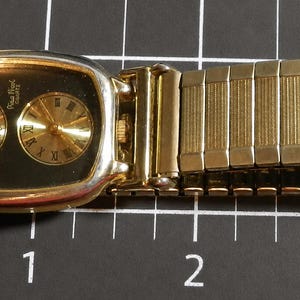 May include: A gold-tone wristwatch with a rectangular case and a black dial featuring two circular sub-dials, one with Arabic numerals and one with Roman numerals. The watch has an expandable link bracelet and measures approximately 3.5 inches long.