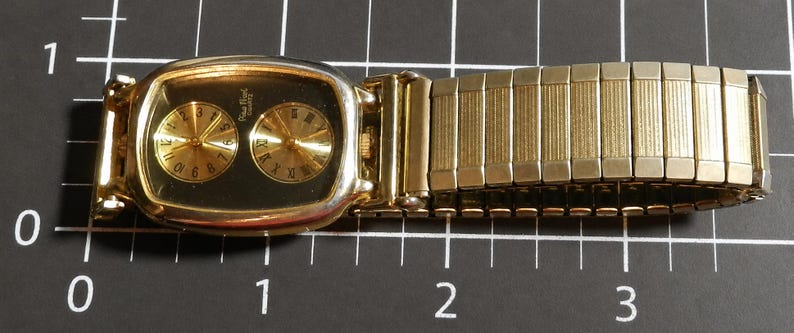 May include: A gold-tone wristwatch with a rectangular case and a black dial featuring two circular sub-dials, one with Arabic numerals and one with Roman numerals. The watch has an expandable link bracelet and measures approximately 3.5 inches long.