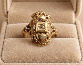 May include: A gold ring featuring an elongated, ornate filigree design with heart-shaped motifs and a central square setting containing a single, small, clear round gemstone. The metalwork displays intricate scrollwork and textured detailing.