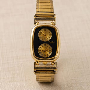 May include: A gold-tone wristwatch with a rectangular case and a flexible expansion band. The black dial features two circular sub-dials with gold faces; the top one has Arabic numerals, and the bottom one has Roman numerals. Paul Picot Quartz is printed.