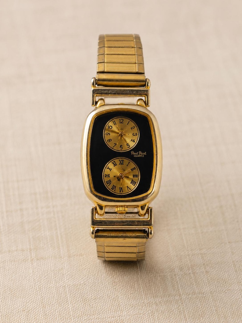 May include: A gold-tone wristwatch with a rectangular case and a flexible expansion band. The black dial features two circular sub-dials with gold faces; the top one has Arabic numerals, and the bottom one has Roman numerals. Paul Picot Quartz is printed.