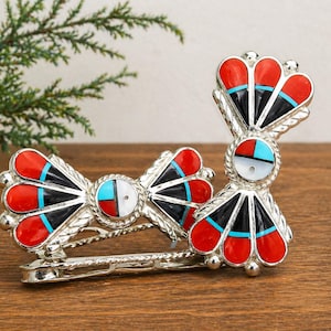 May include: A silver-toned hair barrette with a Southwestern design. The barrette features red, black, and turquoise inlaid stones in a fan-like pattern. The center of the barrette has a white stone with a turquoise accent.