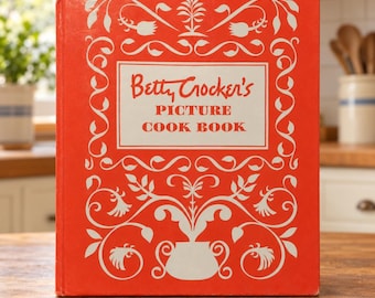 May include: A bright orange hardcover book titled Betty Crocker's Picture Cook Book. The front cover features a cream-colored central rectangle with red text and an intricate white floral and vine pattern surrounding the border and base.