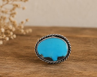 May include: A ring featuring an oval-shaped turquoise stone with a bright blue color and dark matrix inclusions. The stone is set in a polished silver bezel surrounded by a decorative twisted rope metal border, creating a textured frame.