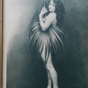 May include: A black and white vintage photograph depicting a figure with long wavy hair, wearing a skirt constructed from large, elongated, pointed leaves that fan out around the waist, creating a voluminous, petal-like silhouette against a dark background.
