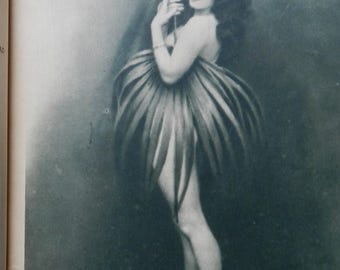 May include: A black and white vintage photograph depicting a figure with long wavy hair, wearing a skirt constructed from large, elongated, pointed leaves that fan out around the waist, creating a voluminous, petal-like silhouette against a dark background.