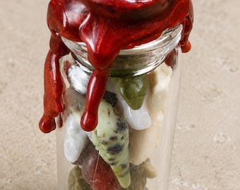 May include: A small glass jar filled with colorful stones, sealed with a vibrant red wax. The wax seal is detailed with an embossed design and drips down the sides of the jar, creating a decorative effect. The stones inside are various shapes and colors.