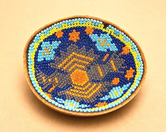 May include: A handcrafted, oval-shaped beaded dish with a wooden rim. The intricate beadwork features a central orange sunburst design, surrounded by blue, yellow, and gray patterns. The dish is a decorative item.