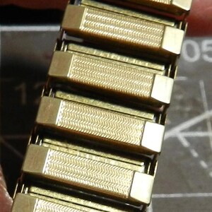 May include: A gold-tone metal expansion watch band featuring a series of rectangular links. Each link has a textured, herringbone-style pattern in the center with smooth, polished end caps. The band is designed to stretch for a comfortable fit.