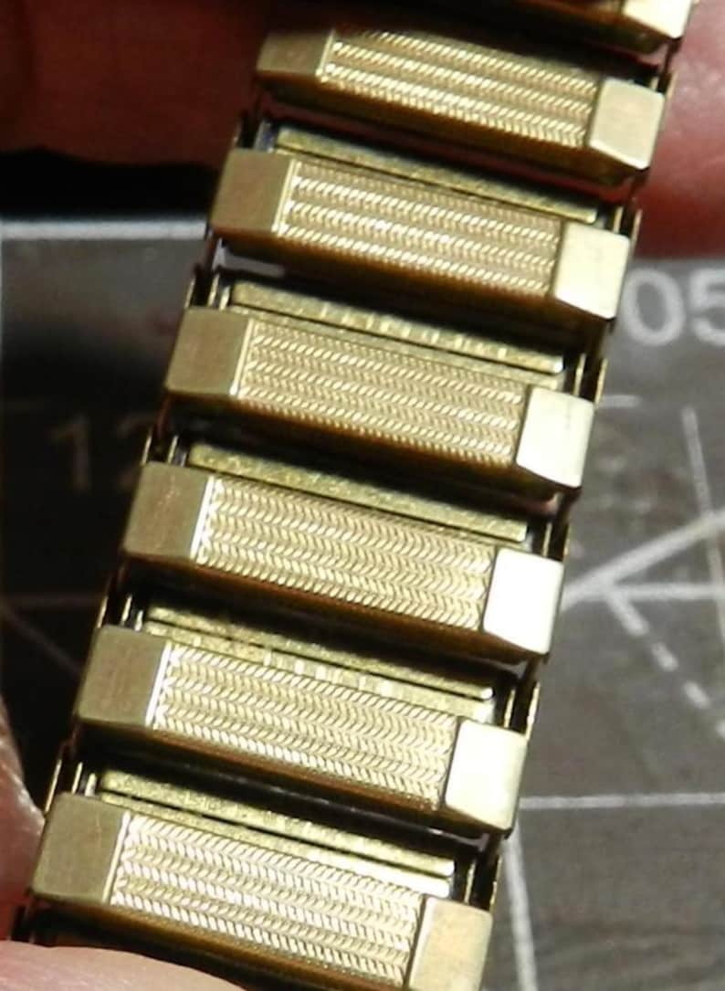 May include: A gold-tone metal expansion watch band featuring a series of rectangular links. Each link has a textured, herringbone-style pattern in the center with smooth, polished end caps. The band is designed to stretch for a comfortable fit.