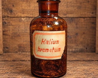 May include: An amber glass apothecary bottle with a wide neck and a matching glass stopper. A rectangular off-white paper label is adhered to the front, featuring the red printed text Kalium bromatum in a distinct, angular, vintage-style font.