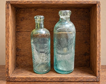 May include: Two vintage light blue glass bottles with embossed lettering. The smaller bottle features the text Anodyne, while the larger bottle displays partial embossed text. Both bottles have rounded shoulders and short, narrow necks with thick rims.
