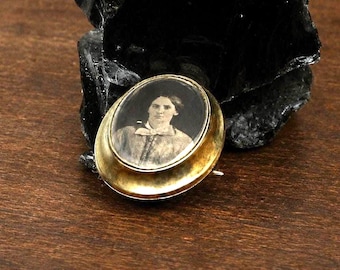 May include: An oval-shaped gold-toned brooch featuring a black and white portrait of a person with dark hair wearing a collared shirt with a bow tie. The brooch has a smooth, polished metallic frame surrounding the central photographic image.