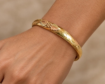 May include: A hinged bangle bracelet crafted from yellow gold, featuring a decorative motif of two rose gold leaves and a small cluster of gold beads at the center. The surface of the metal band displays a textured finish with subtle engravings.