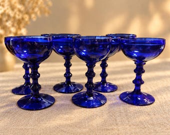 May include: A set of five cobalt blue glass goblets, each featuring a wide, shallow bowl and a stem with a distinct central spherical node, resting on a circular base. The glassware is uniform in color and design, showcasing a deep blue hue.