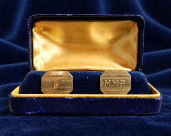 May include: A pair of rectangular gold-toned cufflinks with horizontal line textures, presented in a navy blue velvet box with a gold satin lining. One cufflink features an engraved monogram with the letters M.M.P., while the other is plain.