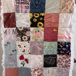 May include: A patchwork quilt made from various fabrics, including shirts with text and patterns. The quilt features squares of different colors and designs, with a white lace border. Some squares have text like "LEVEL I HUMAN" and "READ THIS".