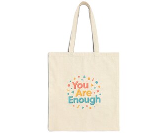 Peut inclure: Sac fourre-tout crème avec un motif coloré. Le motif présente les mots "You Are Enough" dans une police ludique, entourés de points et de formes dans des tons orange, jaune et bleu. Le sac a deux poignées pour le transport.