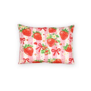 May include: Rectangular pillow with a repeating pattern of red strawberries, white flowers, and red bows on a pink and white striped background. The pillow has a soft, inviting appearance, perfect for adding a touch of whimsy to any room.