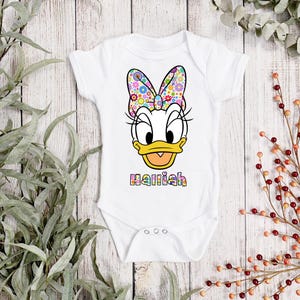 Könnte beinhalten: Weißer Baby-Body mit einem farbenfrohen Daisy Duck-Motiv. Das Design umfasst eine Blumen-Schleife, Wimpern und den Namen "Harriah" in Regenbogenfarben. Der Body hat kurze Ärmel und Druckknöpfe.