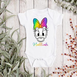 Könnte beinhalten: Weißer Baby-Body mit einem Cartoon-Gesicht von Daisy Duck, einer Regenbogen-Schleife und Wimpern. Der Name "Halliah" steht in Regenbogenfarben darunter. Der Body hat drei Druckknöpfe am unteren Ende.
