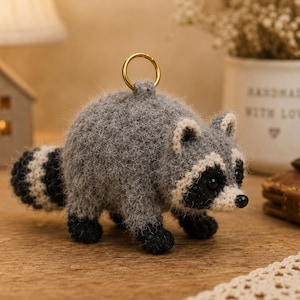 May include: A small, fuzzy, crocheted raccoon keyring featuring a round grey body, a striped tail with black and white bands, black paws, and a white face mask around dark eyes. A gold-toned metal ring is attached to the top of the raccoon.