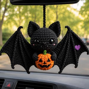 May include: A black crocheted bat hanging ornament with large black eyes, small white fangs, and pink cheek accents. The bat holds a small orange crocheted jack-o'-lantern pumpkin and features a small purple heart embroidered on its right wing.