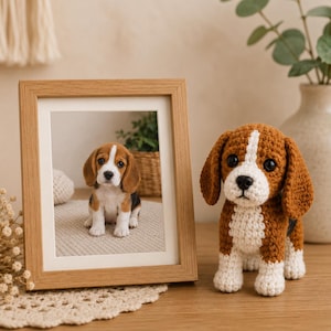 Könnte beinhalten: Eine hellbraun-weiß gehäkelte Beagle-Hundefigur mit schwarzen Augen und einer schwarzen Nase, platziert neben einem hellen Holzbilderrahmen, der ein Foto eines Beagle-Welpen auf einer strukturierten Oberfläche enthält.