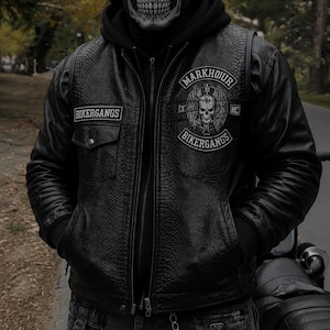 Pode incluir: Um colete de couro preto com acabamento texturizado, apresentando um emblema de caveira com o texto MARKHOUR BIKERGANGS e um emblema BIKERGANGS no peito. O colete é usado sobre um moletom preto, com calças jeans escuras, uma máscara de caveira e um capacete preto.