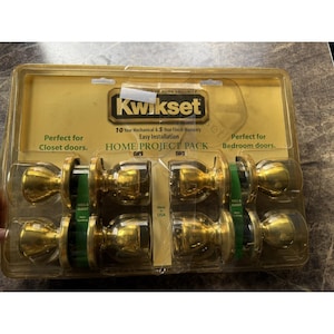 May include: A Home Project Pack of Kwikset door knobs. The package includes six gold-colored door knobs. The packaging is clear plastic with a gold-colored cardboard backing. Text on the packaging includes "10 Year Mechanical & 5 Year Finish Warranty."
