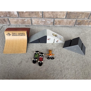 May include: A fingerboard skate park set featuring a tan Tony Hawk Foundation quarter pipe, a gray and white wedge ramp with graphic stickers, a black wedge ramp, two miniature skateboards with graphic decks, and a small orange plastic figure.