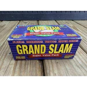 May include: A closed box of "Grand Slam" baseball cards. The box is blue with yellow and red text. The box includes the logos of Fleer, Donruss, Topps, and Upper Deck. The box is on a wooden surface.