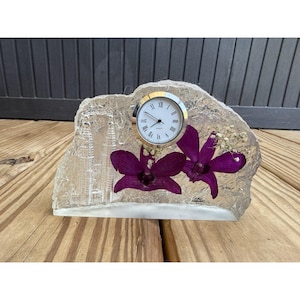 May include: A clear, textured resin desk clock featuring two deep purple orchid blossoms and a small sprig of baby's breath embedded inside. The clock face is round with a gold rim, black hands, and Roman numerals. A faint etched image of towers is visible.
