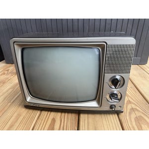 May include: A vintage Quasar television set featuring a black and silver plastic casing. The front panel includes a screen, a speaker grille with a grid pattern, two large circular channel tuning knobs, and a smaller power button at the bottom.