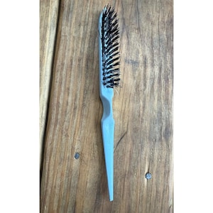 Könnte beinhalten: Eine hellblaue Haarbürste mit schwarzen Borsten. Die Bürste hat einen langen, schlanken Griff und einen rechteckigen Kopf mit eng stehenden Borsten. Die Bürste ist aus Kunststoff und dient zum Stylen der Haare.