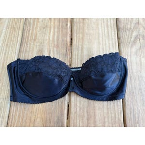 May include: A black strapless bra with lace detailing across the top. The bra has a smooth, satin-like finish and a small decorative element between the cups. The bra is displayed on a wooden surface.