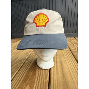 May include: A baseball cap featuring a beige crown and a dark gray brim. The front of the crown displays a prominent embroidered patch of a yellow scallop shell with a red outline. The cap is constructed with six panels and a stitched brim.