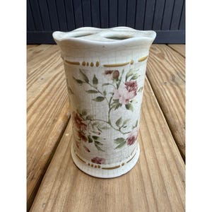 May include: A cream-colored ceramic toothbrush holder with a floral design. The holder features pink roses and green leaves on a textured background. Gold accents are present around the top and bottom.