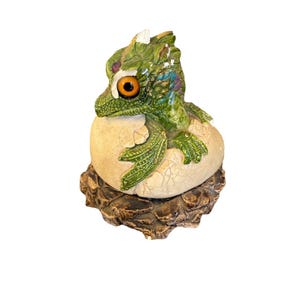Peut inclure: Figurine de dragon vert et jaune éclos d'un œuf fissuré. Le dragon a un œil orange et est perché sur une base marron texturée. L'œuf est de couleur crème.