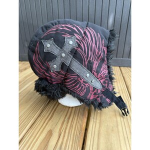 May include: A black trapper hat featuring a dark pink graphic pattern of wings and a prominent grey cross applique with silver-toned metal studs. The hat is lined with black faux fur and includes a black adjustable chin strap with a plastic buckle.