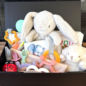 May include: A black gift box filled with baby items. Includes a gray plush bunny, a plush sheep, a book with an elephant illustration, and various baby toys in shades of pink, orange, and green.
