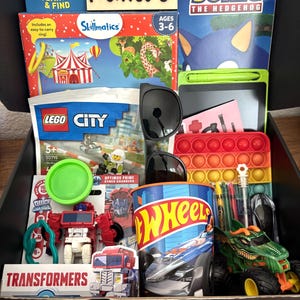 May include: A black box filled with toys and games, including a Sonic the Hedgehog book, a Lego City set, a Hot Wheels cup, and a Transformers toy. The box also contains a green drawing tablet, sunglasses, and a pop-it toy. The box is labeled "created by Huxley."