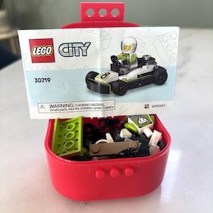 Puede incluir: Un contenedor de almacenamiento LEGO City rojo con un coche de carreras parcialmente ensamblado y una hoja de instrucciones impresa. La hoja muestra un coche de carreras con un conductor. El contenedor est&aacute; lleno de varias piezas de LEGO.